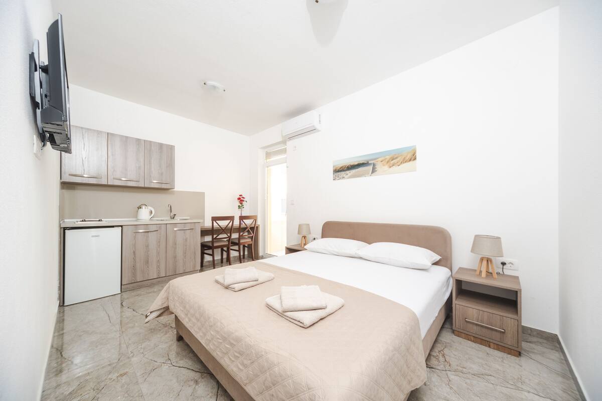 A hotel room with a bed, a desk, a chair, a TV mounted on the wall, and a small table with a lamp.