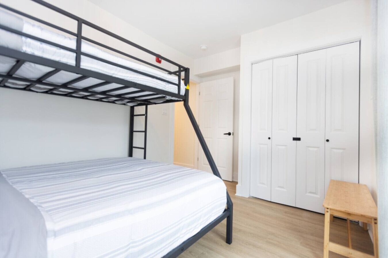 Una cama litera con escalera, un taburete de madera y un armario blanco en una habitación con suelo de madera.