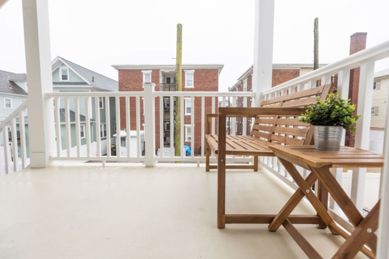 A balcony with a wooden bench and a potted plant, white railings, and a view of neighboring houses.