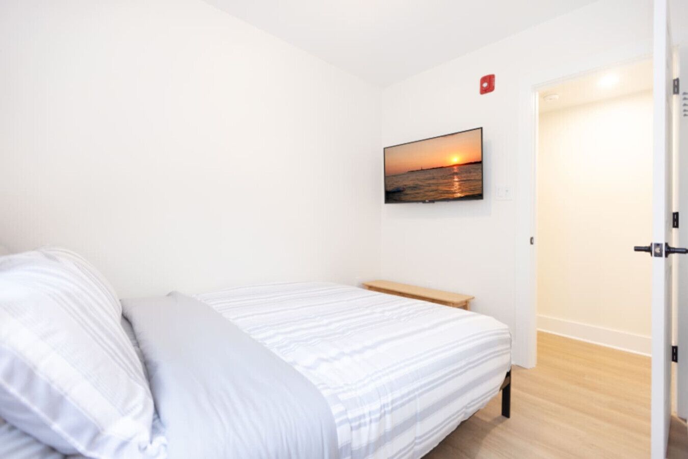 A bedroom with a bed, a wooden bench, a wall-mounted TV, and a door.