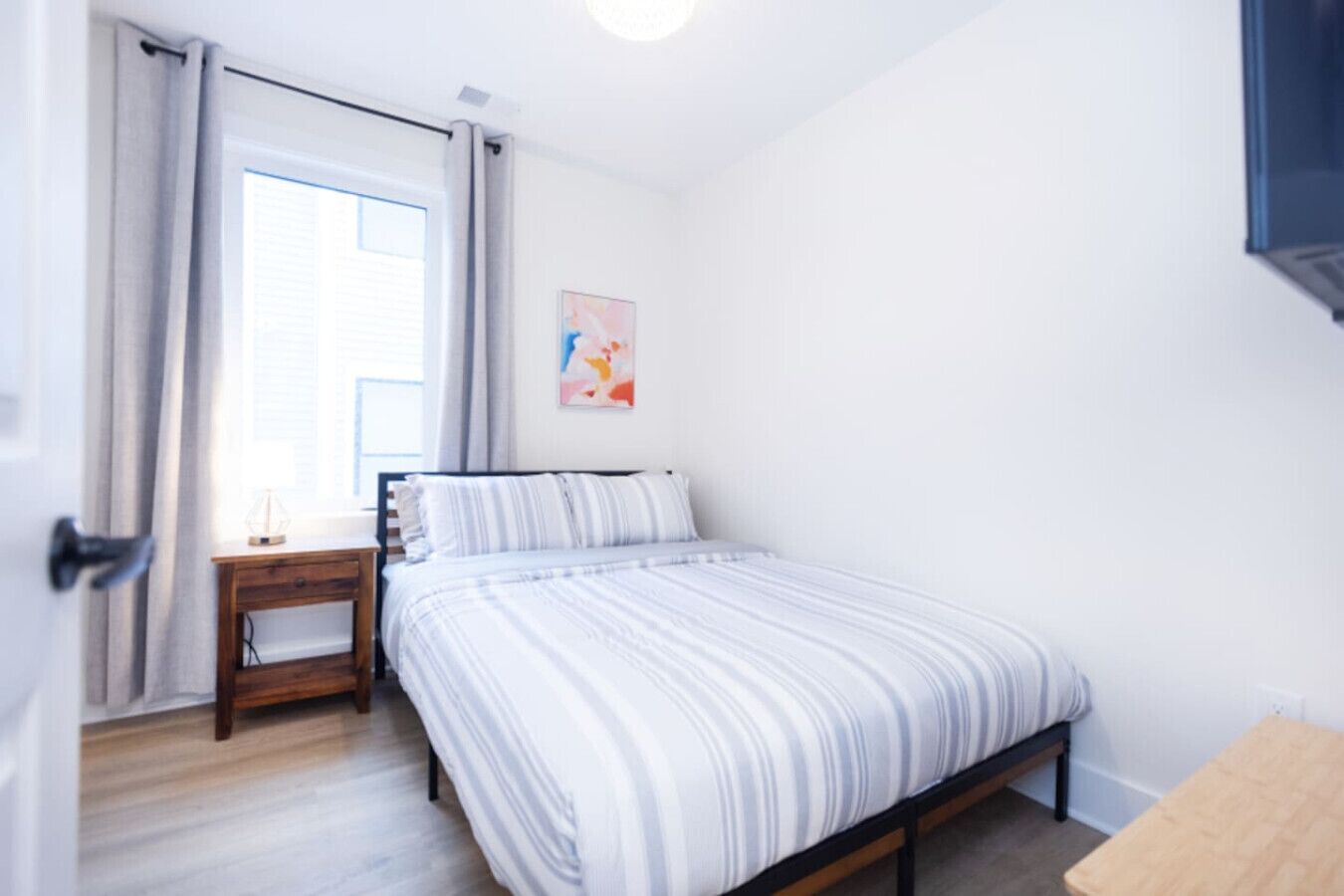 Een slaapkamer met een bed, een nachtkastje, een raam met gordijnen en een kunstwerk aan de muur.