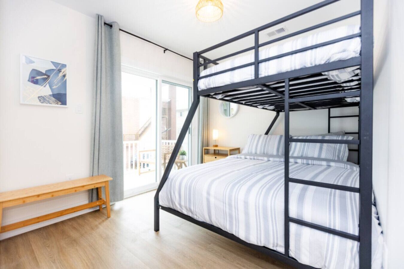 A bunk bed with a striped bedspread, a wooden bench, a bedside table with a lamp, and a window with a view of buildings.