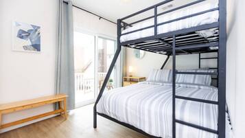 A bunk bed with a striped bedspread, a wooden bench, a bedside table with a lamp, and a window with a view of buildings.