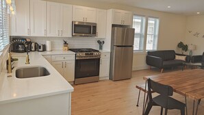 A modern kitchen with white cabinets, stainless steel appliances, a wooden dining table, and a black sofa. - Steps to THE Beach – 3 BDR Condo + Parking (Wildwood)