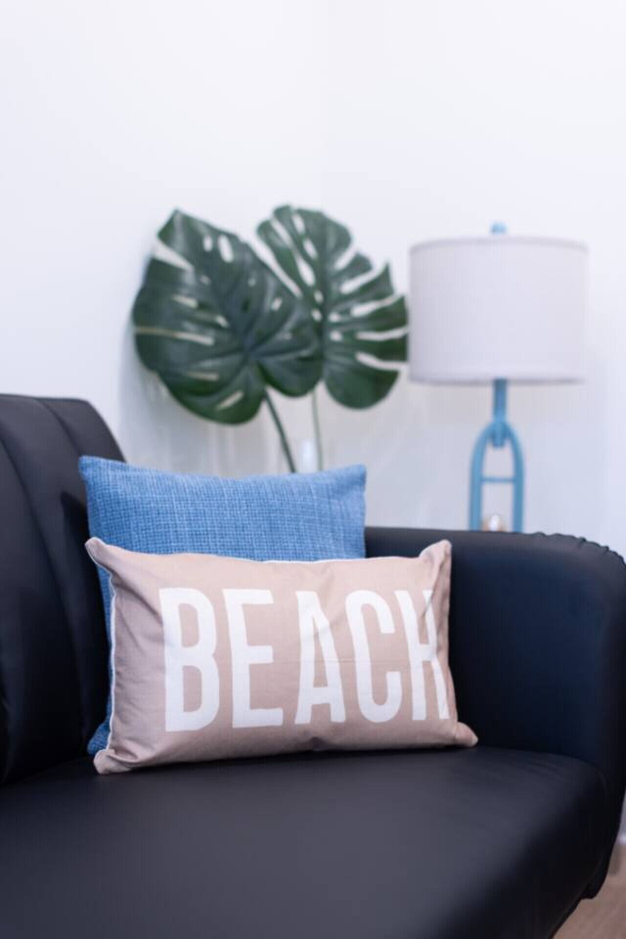 A black sofa with a beige pillow that reads "BEACH" and a blue pillow.