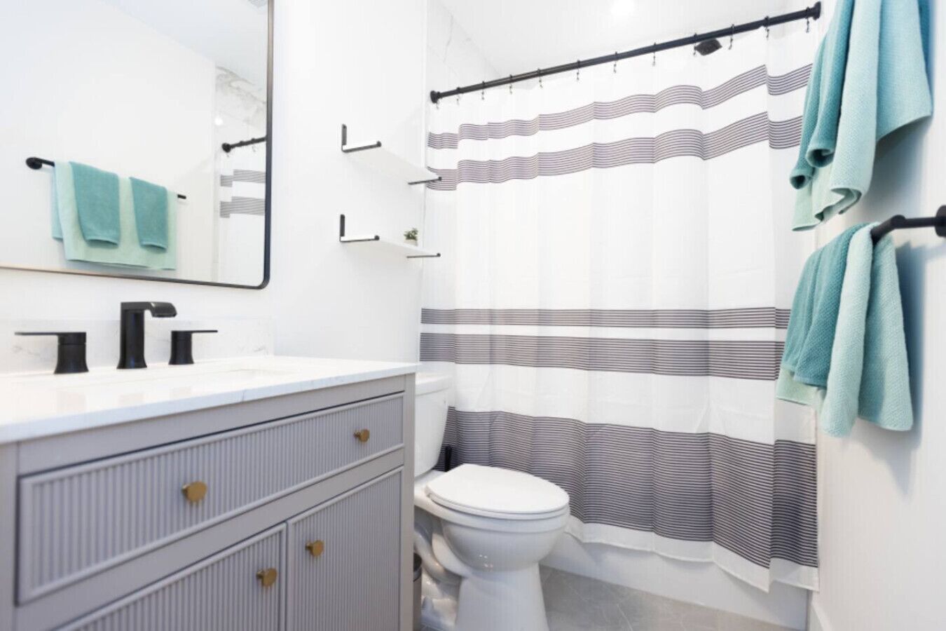 Une salle de bain avec un lavabo blanc, un robinet noir et des toilettes. On y trouve un rideau de douche à rayures et un porte-serviettes avec des serviettes.