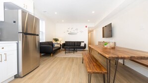 A modern kitchen with a stainless steel refrigerator, a wooden dining table with benches, a black sofa, and a wall-mounted TV. - Amazing all new 1fl condo> Steps to The Beach (Wildwood)
