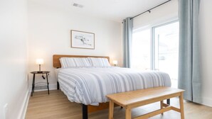 A bedroom with a bed, bedside tables, a bench, and a large window with curtains. - Amazing all new 1fl condo> Steps to The Beach (Wildwood)