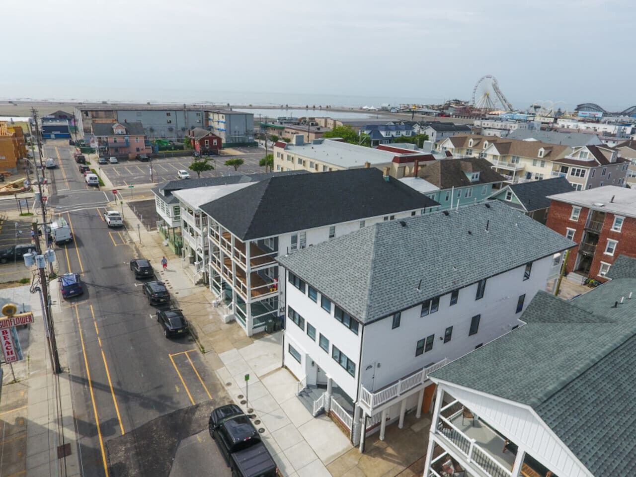 A coastal town with a prominent white building with a grey roof, a parking lot, and a view of the sea and a large Ferris wheel in the distance.