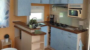 A compact kitchen with blue cabinets, a microwave, a coffee maker, and a small dining area with a table and chairs. - Spacious mobile home with terrace and lake view, ideal for families. (Bairon et ses environs)