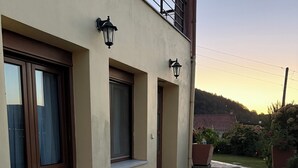 Un bâtiment doté de trois fenêtres, chacune ayant un petit rebord et une porte. Deux lampes sont fixées au mur au-dessus des fenêtres. Le bâtiment est entouré d’une cour pavée de pierres, et un objet sphérique jaune se trouve sur le sol.