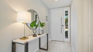 A hallway with a white console table, a floor lamp, two potted plants, a mirror, and a door leading to another room.