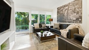 A modern living room with a large window, a black leather sofa, a coffee table, and a cityscape painting on the wall.