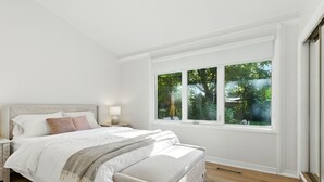 A bedroom with a large window, a bed, a bedside table, and a nightstand.