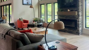 A spacious living room with a large TV, a stone fireplace, and large windows. - Newly constructed, beautiful, modern lakehouse with new barrel sauna  (Mactier)