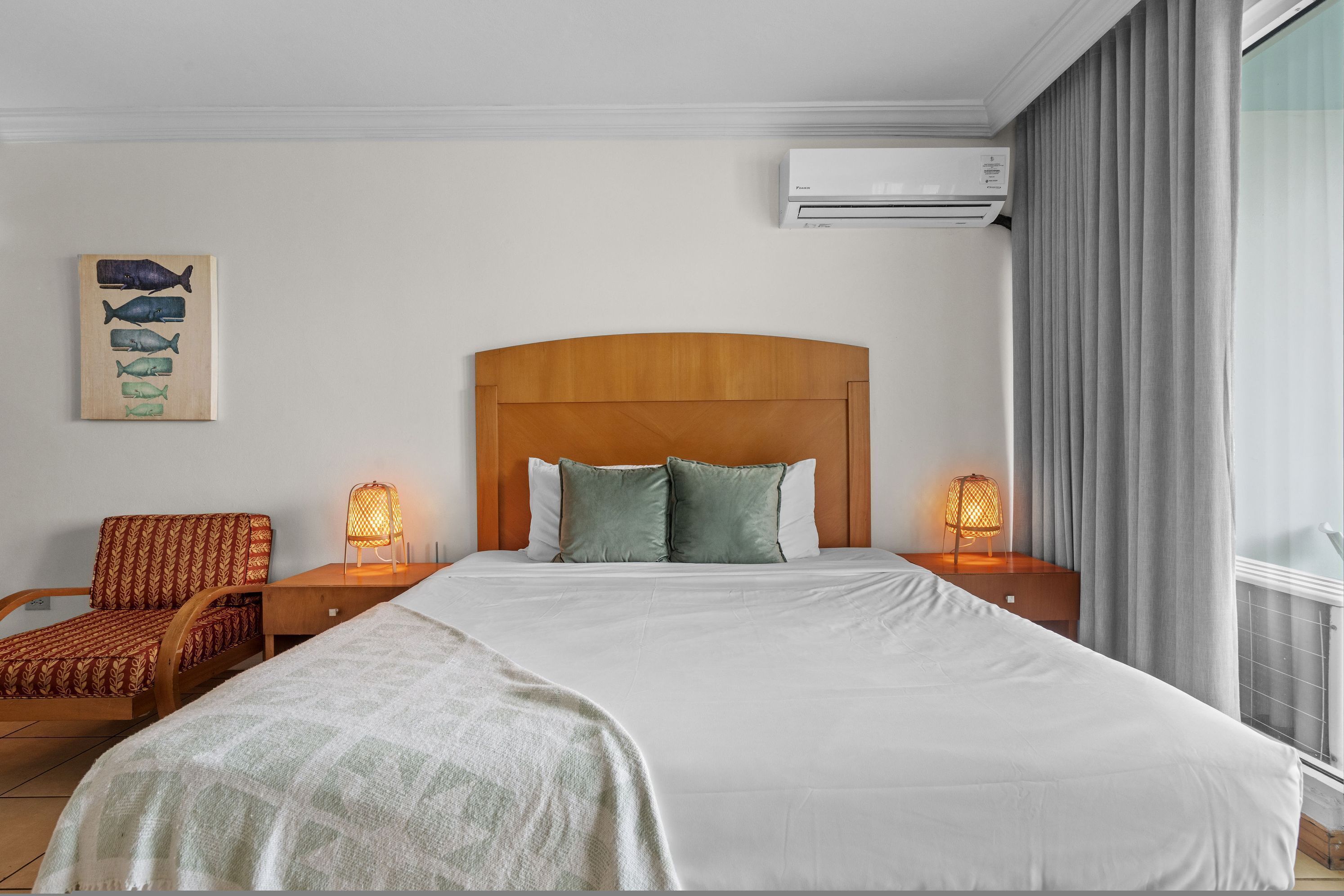 Ein Hotelzimmer mit massivem Holzkopfteil, einem Bett mit weißer Bettwäsche, zwei Nachttischlampen, einem Sessel und einer Wandklimaanlage.
