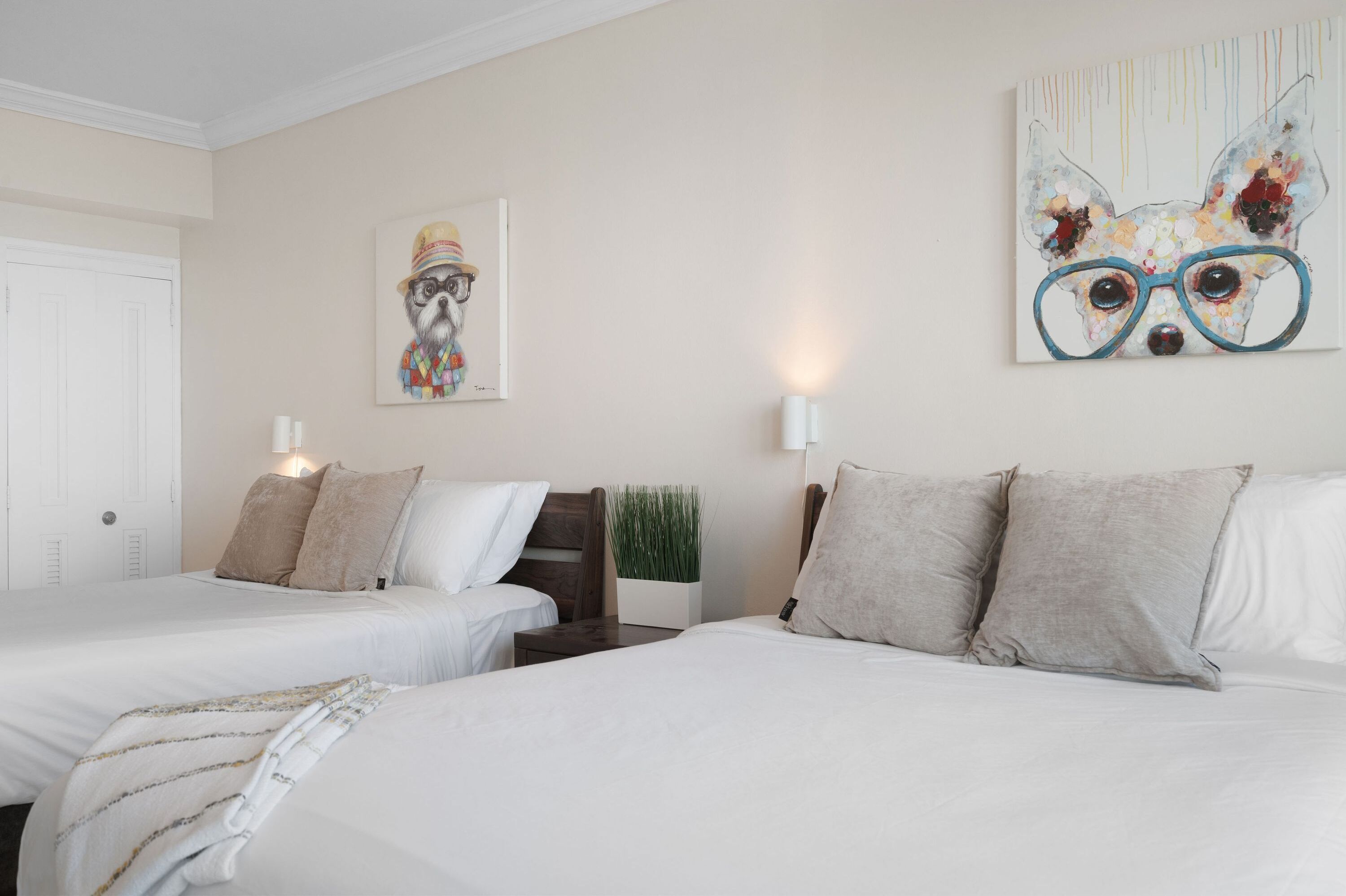 A hotel room with two beds, a nightstand, a wall-mounted lamp, and a painting of a dog wearing glasses.