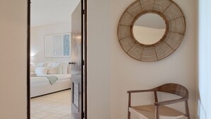 An open door leading to a bedroom with a bed and a large window. A round woven mirror hangs on the wall, and a wooden chair is positioned near the door. - Beachfront Paradise 2BR Crescent Beach Seaside (Humacao)