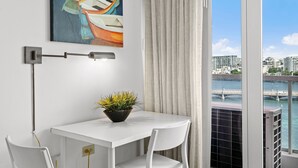 Une chambre moderne avec une table blanche, une plante en pot, un tableau représentant des bateaux et, par la fenêtre, une vue sur un pont et des bâtiments.