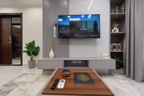 Una sala de estar moderna con un televisor de pantalla plana montado en la pared, una mesa de centro de madera con controles remotos y un libro, y una planta en maceta.