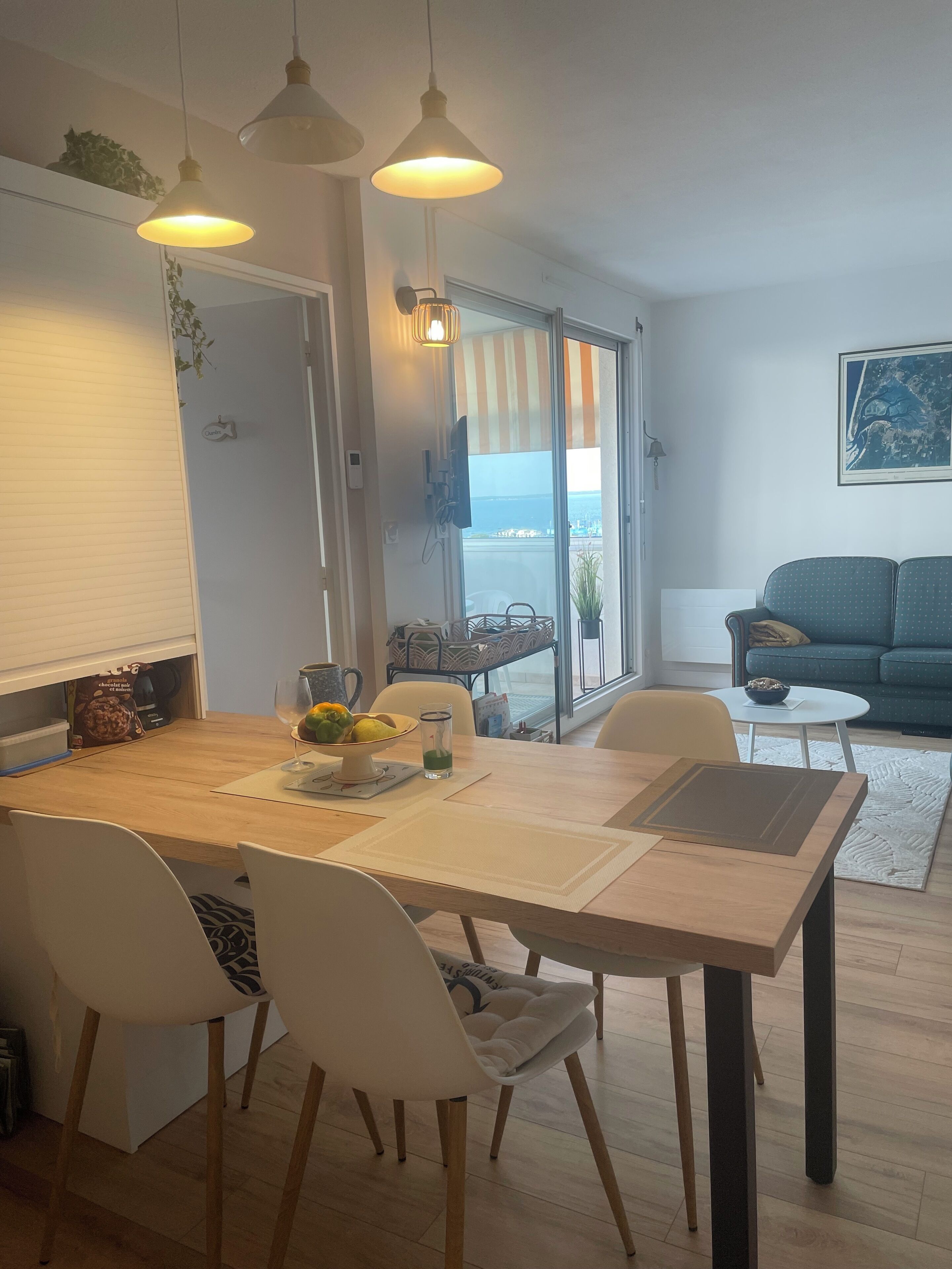 A dining area with a wooden table, white chairs, and pendant lights. A large window offers a view of the outdoors.