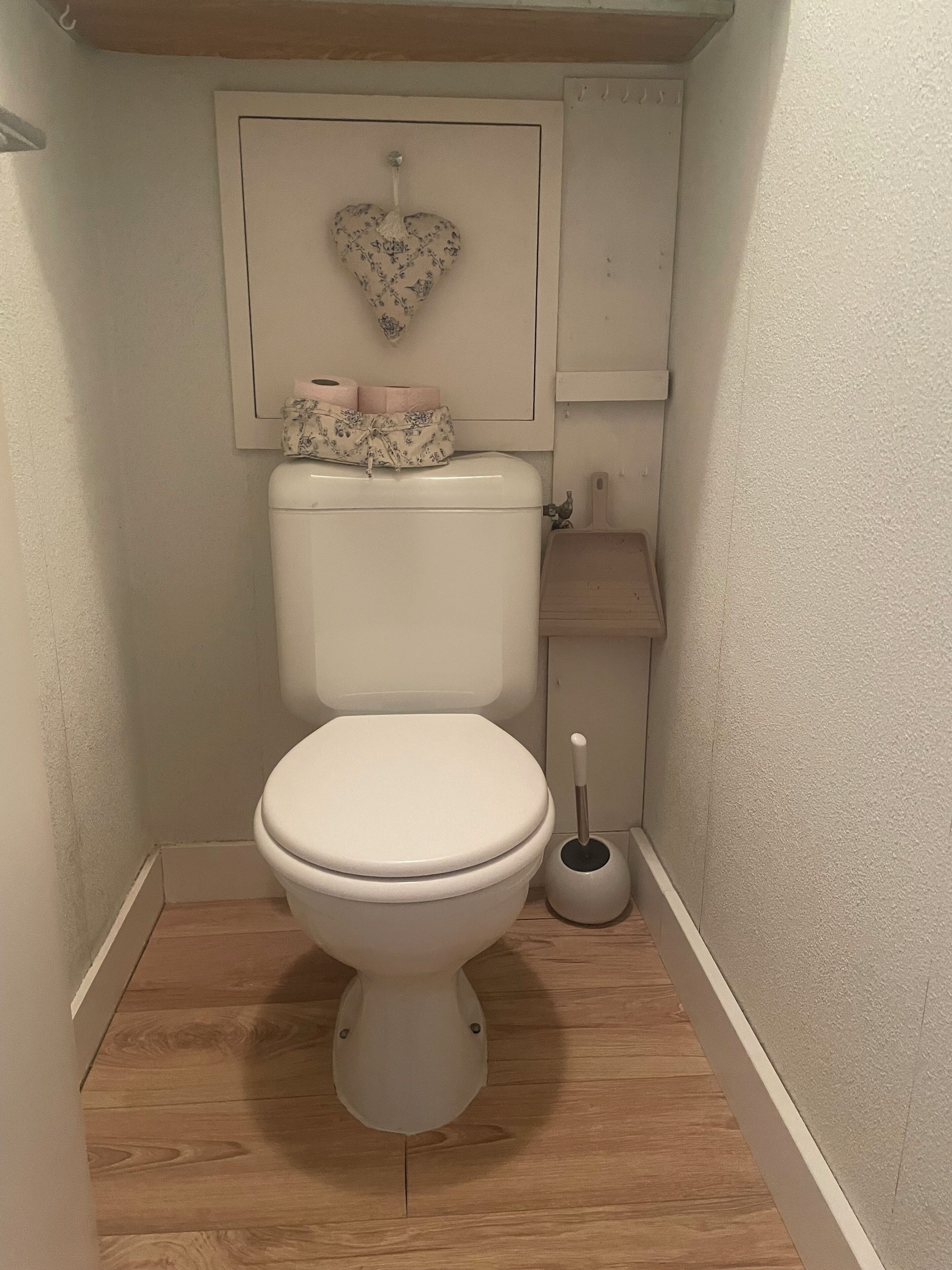 Een kleine badkamer met een toilet, een toiletborstel en een hartvormige decoratie aan de muur.