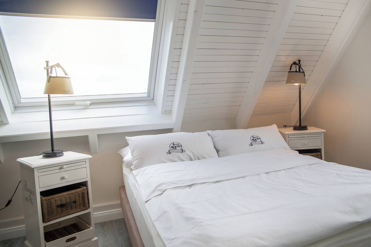 Ein Schlafzimmer mit Dachfenster, zwei Nachttischen mit Lampen, einem Bett mit weißer Bettwäsche und einem kleinen, weißen Beistelltisch mit Lampe.