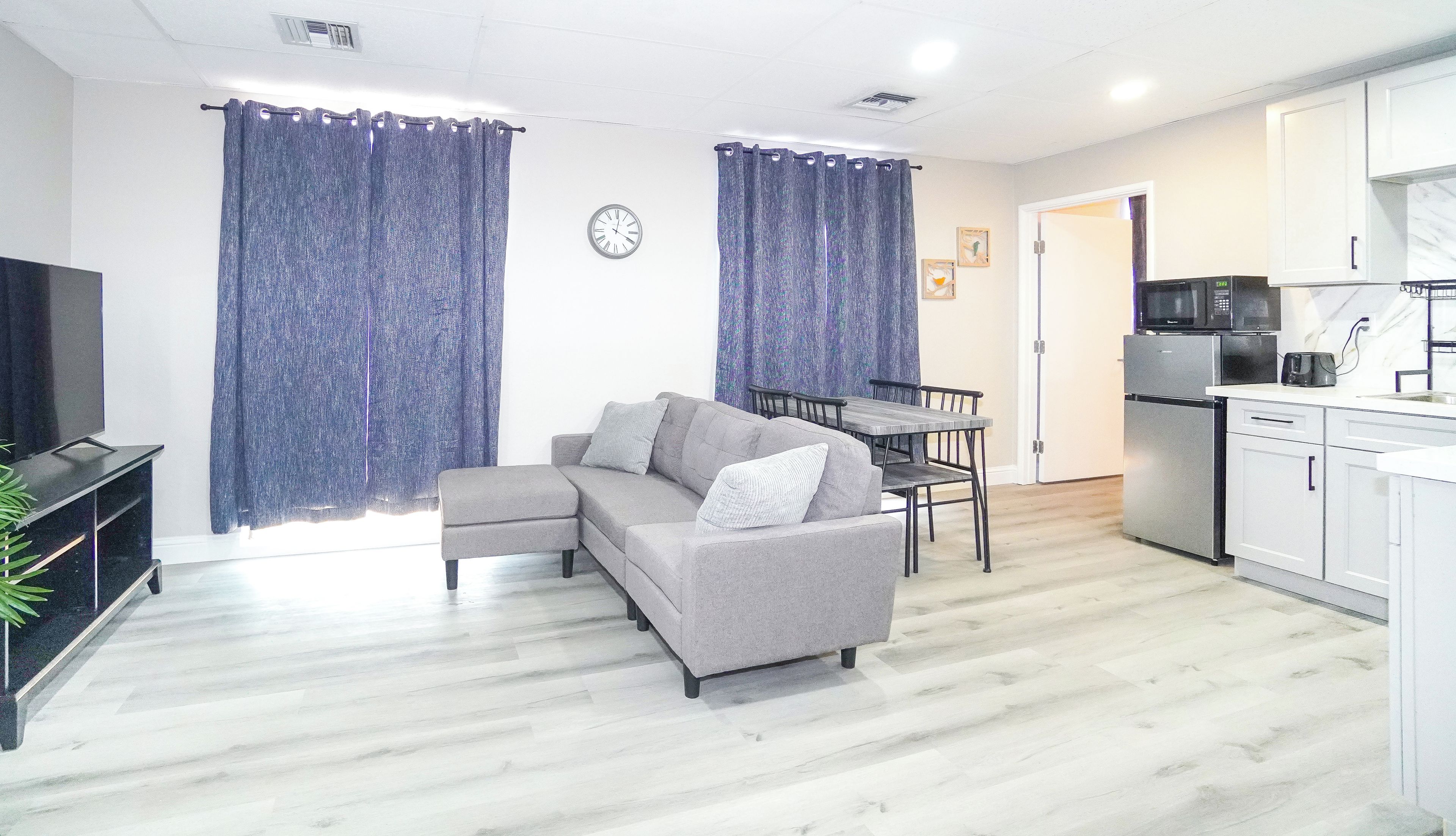 A living room with a grey sofa, a dining table, and a kitchen area with a microwave and refrigerator.