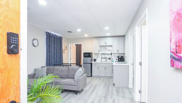Una sala moderna con un sofá gris, una mesita de centro y una planta en maceta. Se ve una cocina con armarios blancos y un microondas al fondo.