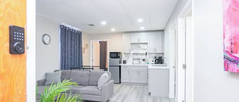 A modern living room with a grey sofa, a coffee table, and a potted plant. A kitchen with white cabinets and a microwave is visible in the background.