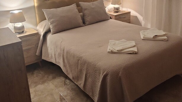 Ein ordentlich bezogenes Bett mit einem gepolsterten Kopfteil, zwei Nachttische mit Lampen und ein gefaltetes Handtuch auf dem Bett.