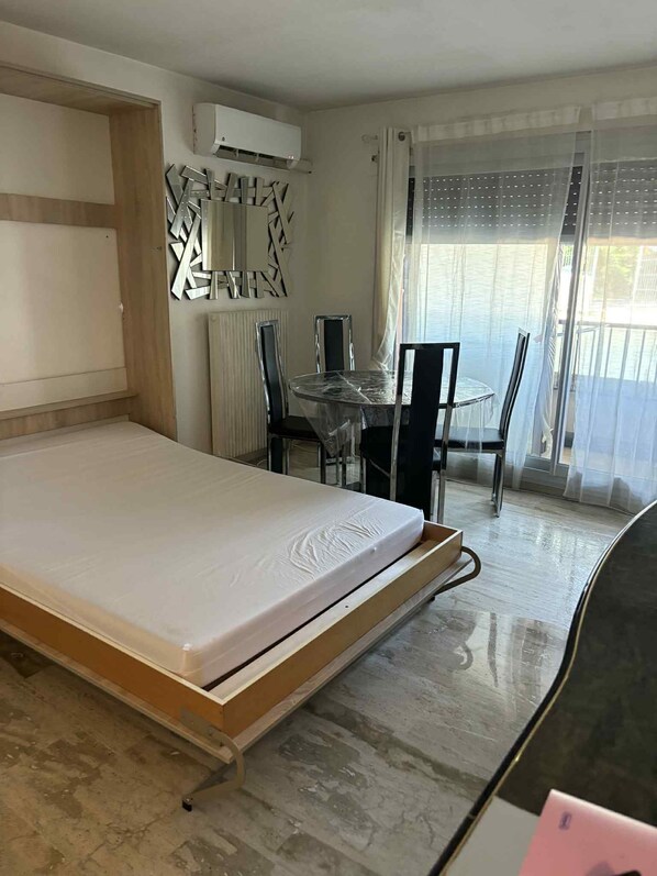 A compact room with a foldable bed, a dining table with chairs, a decorative wall piece, and a window with sheer curtains. - Les 3 rivieres à mandelieu (Mandelieu-La-Napoule)