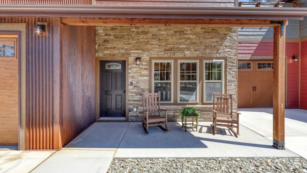 A covered patio area with two wooden chairs and a small table, a stone wall, and a garage door. - Gold Rush Charm Meets Modern Comfort (Rapid City)