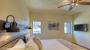 A bedroom with a bed, a ceiling fan, a mounted TV, and two windows. - Gold Rush Charm Meets Modern Comfort (Rapid City)