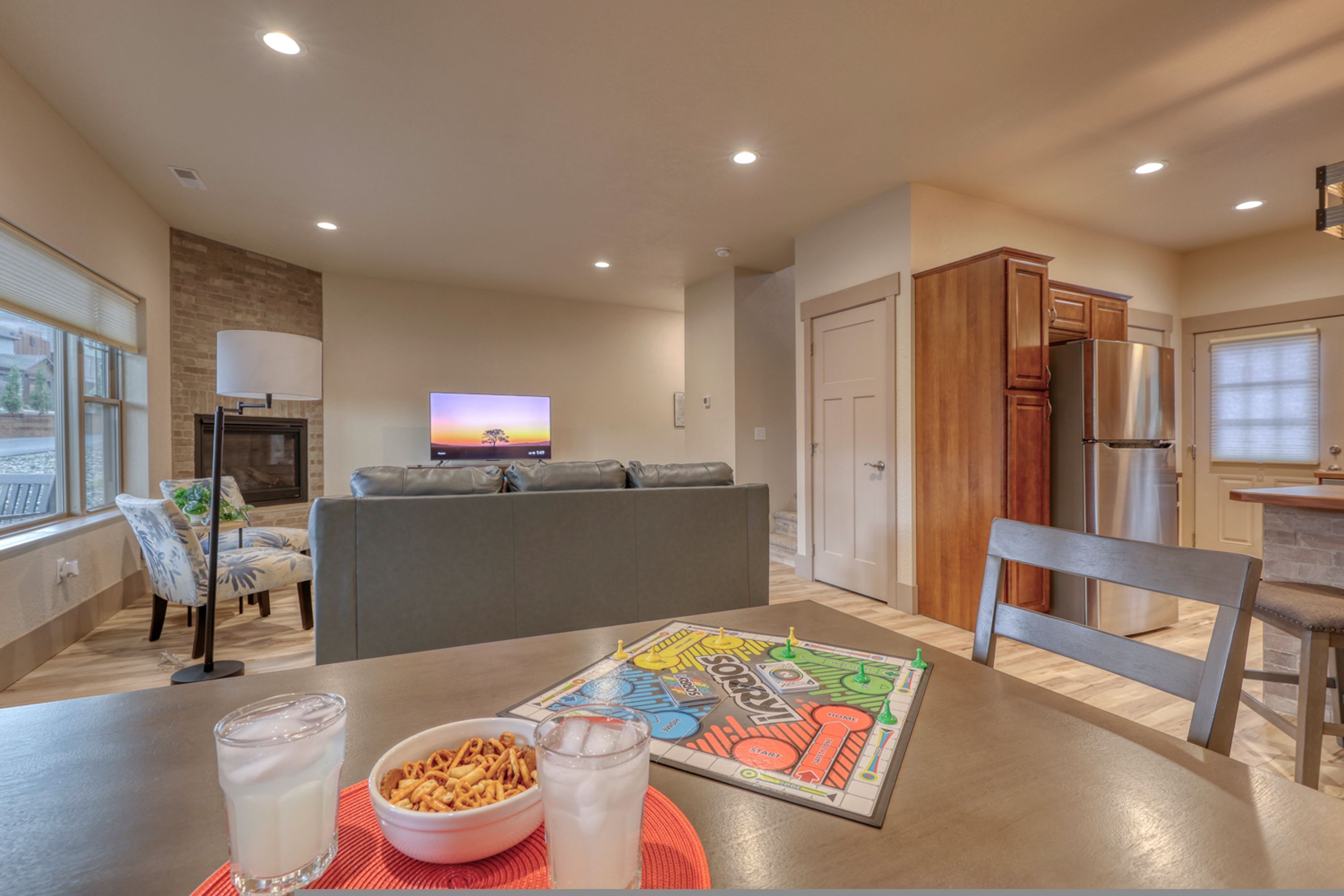 Una sala moderna con un sofá gris, una mesa de centro con bocadillos y bebidas, un televisor mostrando un atardecer, y una zona de cocina con armarios de madera y un refrigerador.