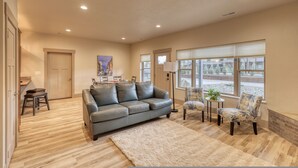 A spacious living room with a grey leather sofa, a coffee table, and a large window with blinds.