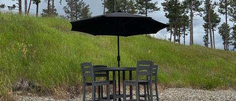 A patio table with four chairs and a large umbrella, set on a gravel surface with a backdrop of tall pine trees.