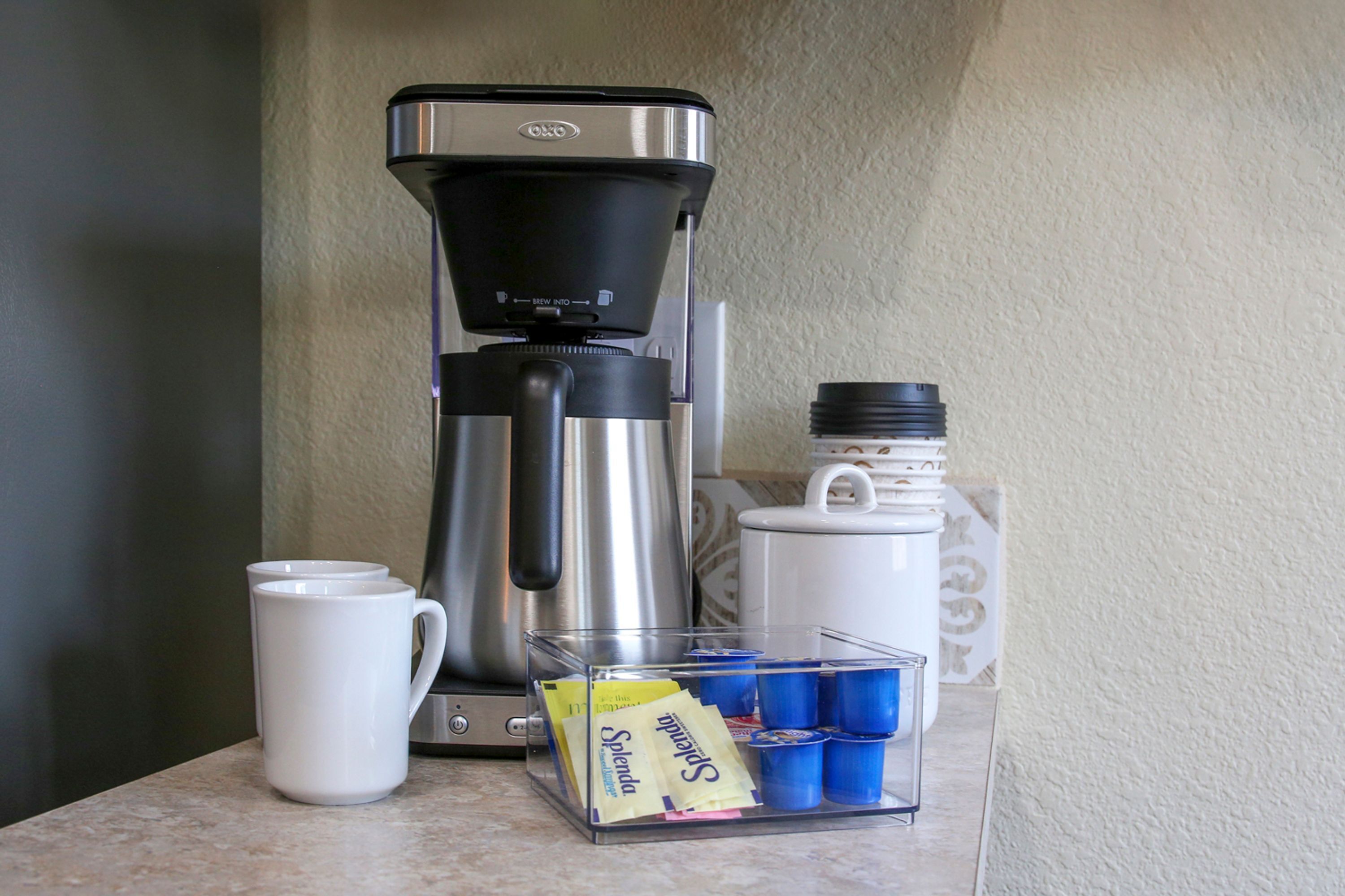 A coffee maker, a white mug, a coffee pot, and a container with coffee cups and a Splenda packet on a countertop.