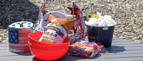 A table with a variety of food items and drinks, including a bucket of ice, a cooler, and a basket with snacks.