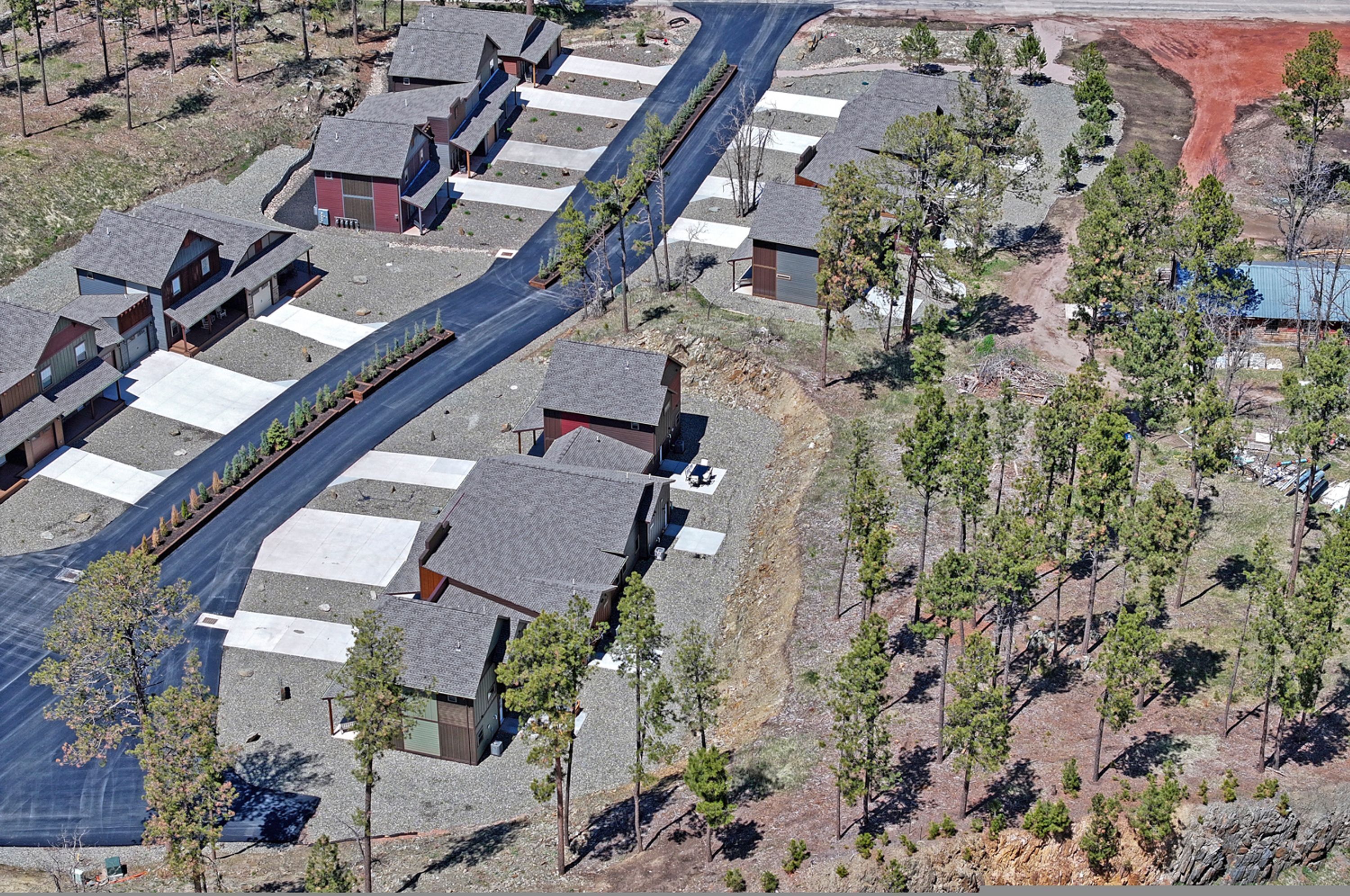 An aerial view of a residential area with houses arranged in a grid pattern, surrounded by trees and a dirt road.