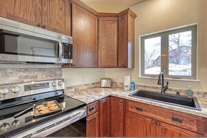 Una cocina con armarios de madera, un microondas de acero inoxidable, un horno, un lavamanos y una ventana con vistas a un paisaje nevado.