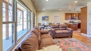 A spacious living area with a large window, a brown leather sofa, a patterned rug, and a kitchen in the background.