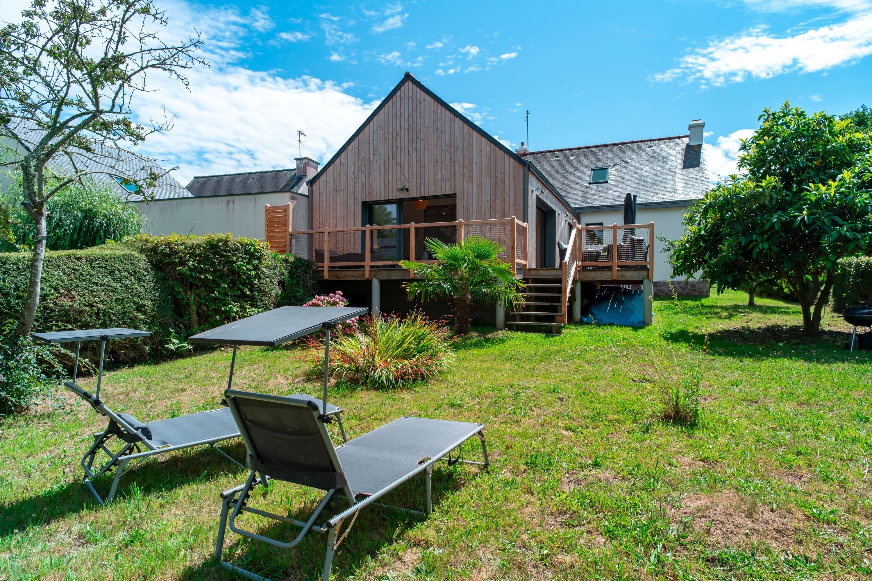 Una casa moderna con fachada de madera, un balcón y un patio trasero con césped y dos sillones de descanso.