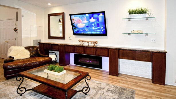 Een woonkamer met een bruine leren bank, een salontafel met een glazen blad, een flatscreen televisie boven een open haard en een kleine plant op een plank.