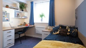 A bedroom with a bed, a desk with a laptop, a chair, a radiator, and a window with curtains. - For Students Only Kimber Student Residence (Southampton)