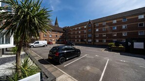 A parking lot with a black car, a white van, and a red car parked. There are buildings with multiple windows and a church spire in the background. - For Students Only Kimber Student Residence (Southampton)