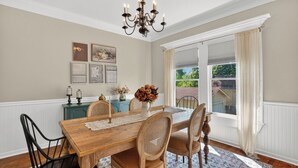 A dining room with a wooden table, chairs, a chandelier, and a view through a window. - 1921 Craftsman Bungalow · Charnwood Historic District (Tyler)