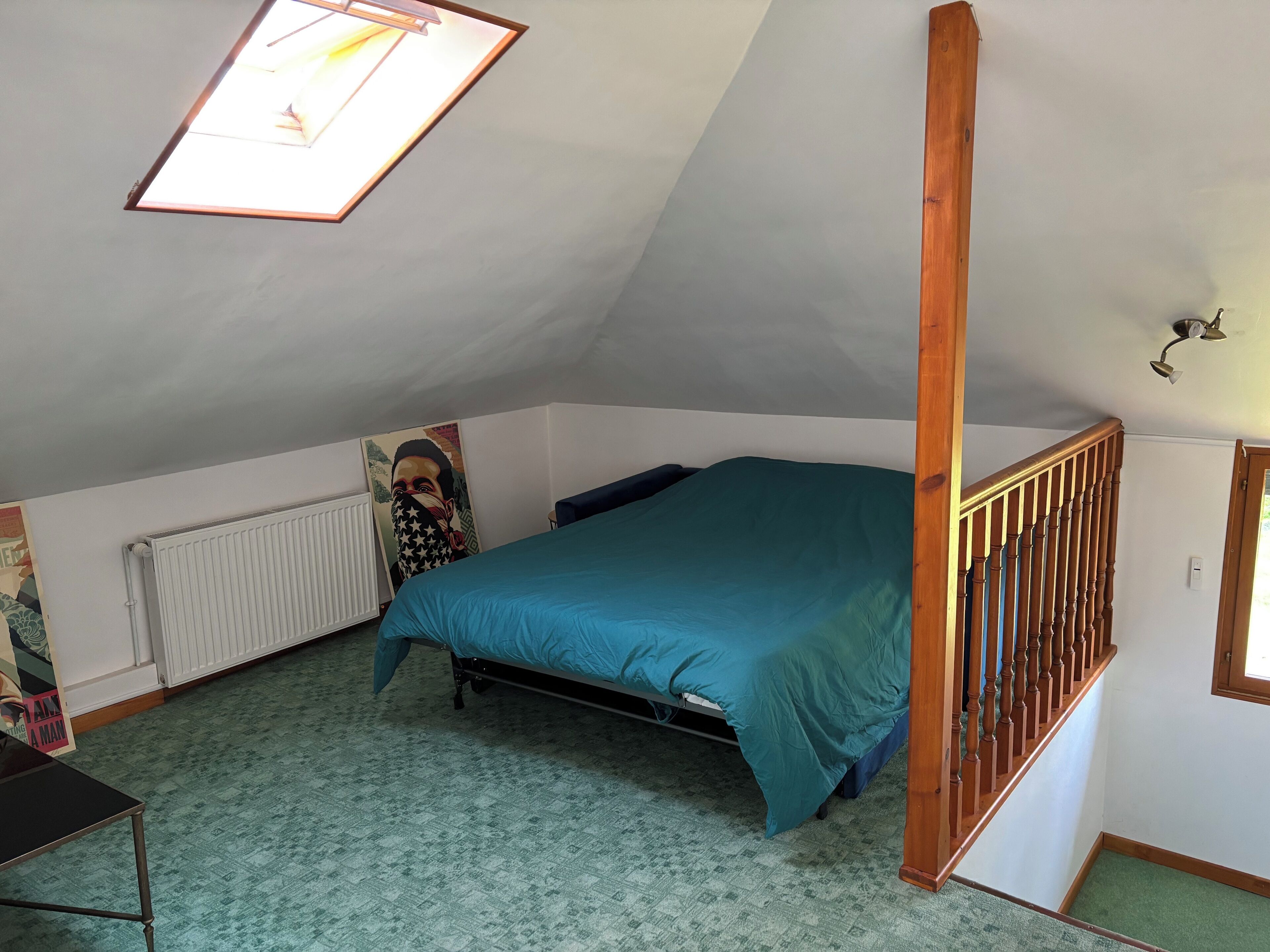 Une chambre avec un plafond en pente, un lit, un radiateur, un escalier en bois et une fenêtre.
