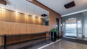 A modern lobby with a wooden panel wall, a black bench, a large potted plant, and a glass elevator. - DRL - Apartments 300m From Bombinhas Beach (Bombinhas)