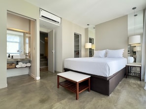A modern hotel room with a large bed, a bedside table, a small bench, a wardrobe, a mirror, and an air conditioning unit.